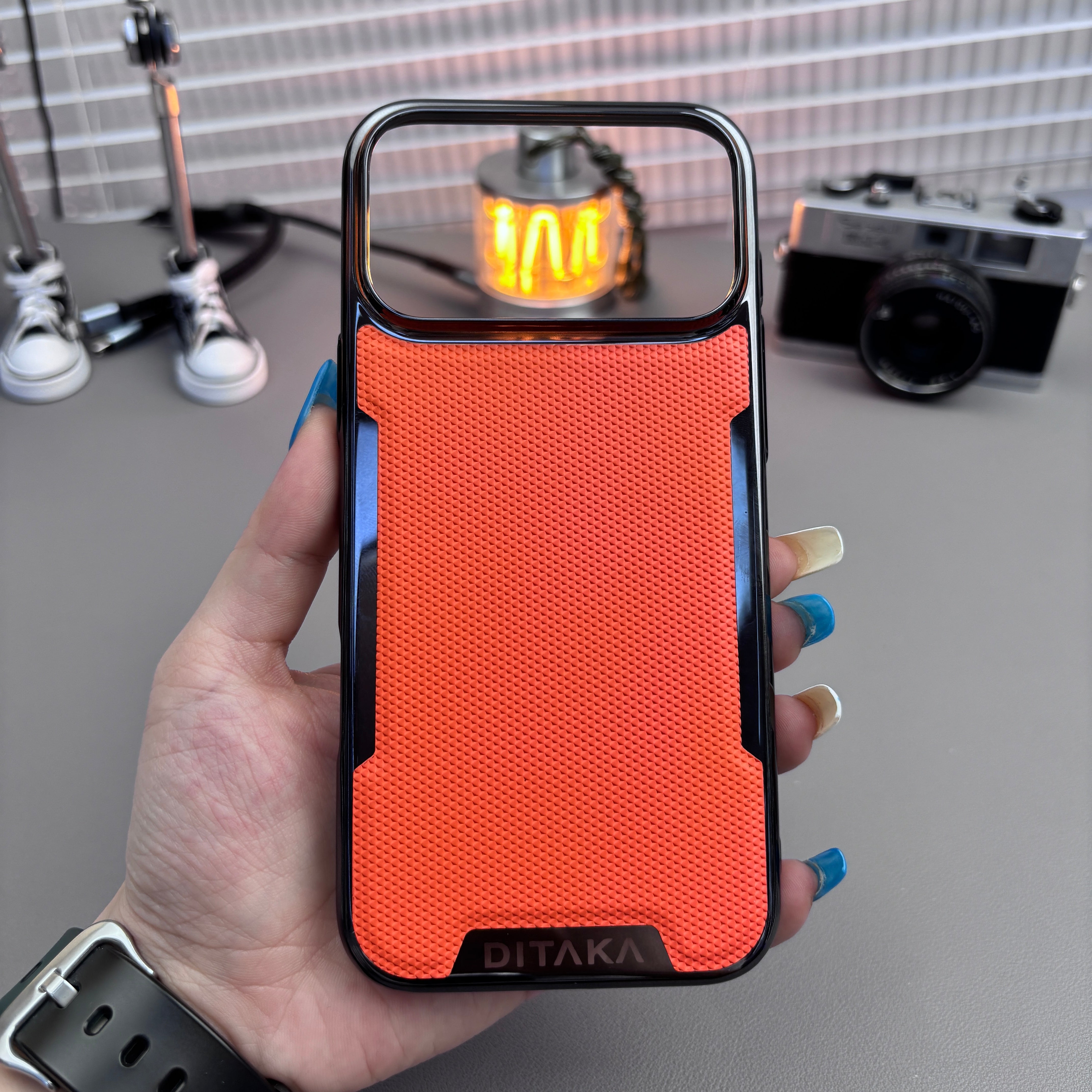 Hand holding a phone with an orange case, with a camera and equipment in the background.