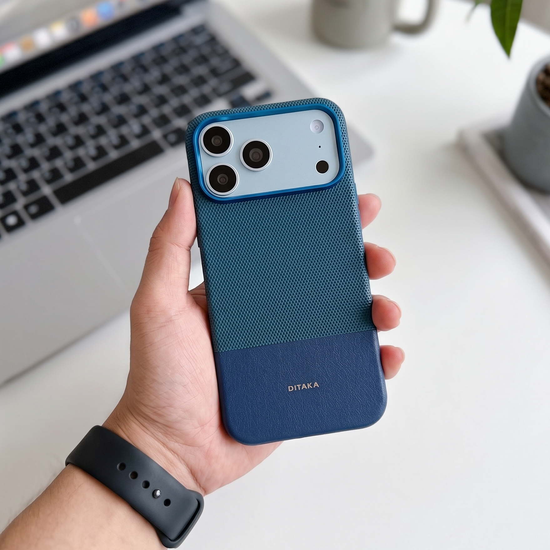 Hand holding a blue phone case with a laptop and desk in the background