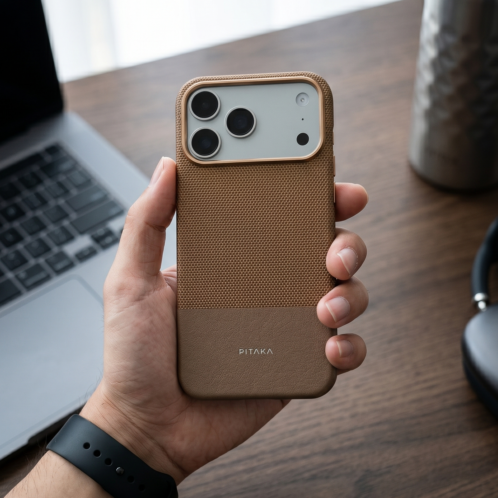 Hand holding a brown phone case with 'PITAKA' branding on a wooden desk with a laptop and headphones.