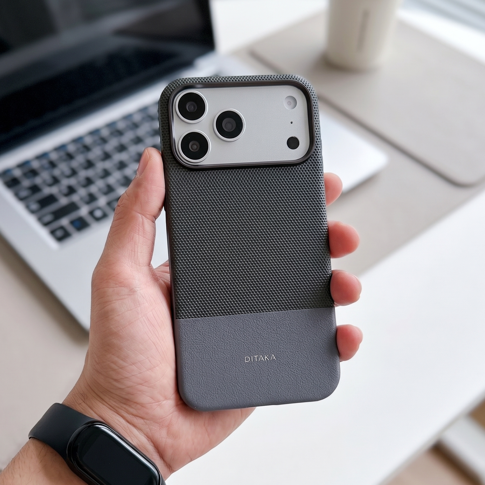 Hand holding a black phone case with a laptop and coffee cup in the background